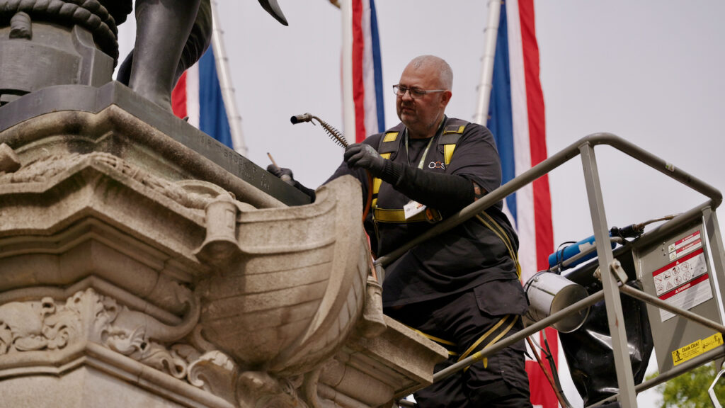 A worker in safety gear stands on a lift, cleaning a large stone statue. Red, white, and blue flags are visible in the background. The worker holds a hose and appears focused on the task.