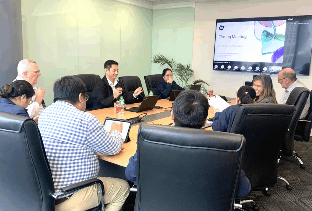 A group of people sit around a conference table in a meeting room, viewing a presentation titled Closing Meeting on a large screen. Some take notes while others engage in discussion.
