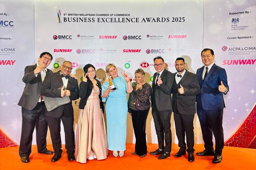 A group of eight people in formal attire stand on a red carpet, smiling and giving thumbs up at the British Malaysian Chamber of Commerce Business Excellence Awards 2025. An awards backdrop with sponsor logos is behind them.