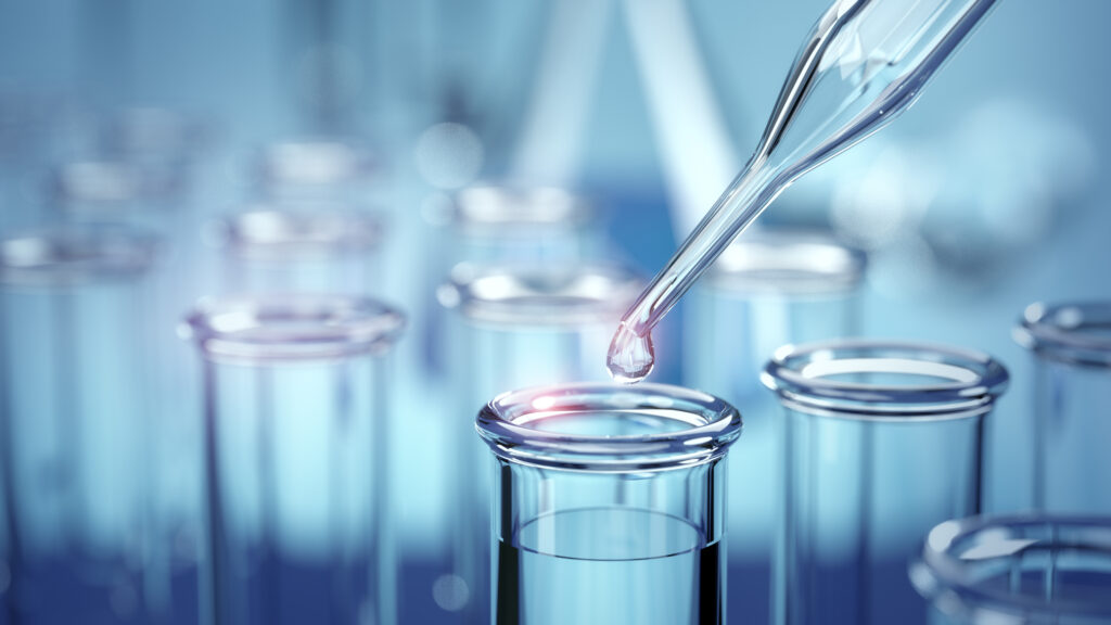 A close-up of a pipette dropping liquid into a glass test tube, with several other test tubes blurred in the background, in a laboratory setting with a blue tone.