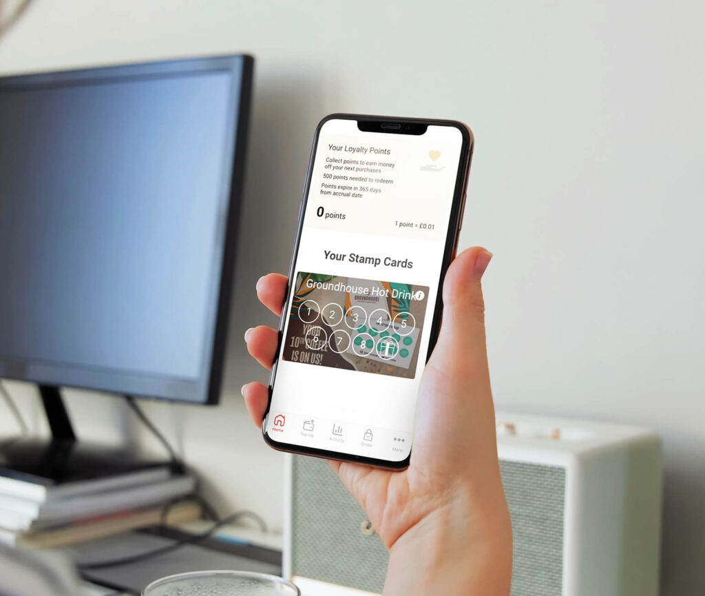 A hand holds a smartphone displaying a loyalty points and stamp card app, with a computer monitor, keyboard, and speaker visible in the background on a desk.