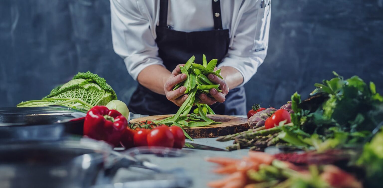 A chef wearing a white coat and dark apron prepares fresh green snap peas at a kitchen counter surrounded by assorted vegetables, including cabbage, peppers, carrots, and leafy greens.