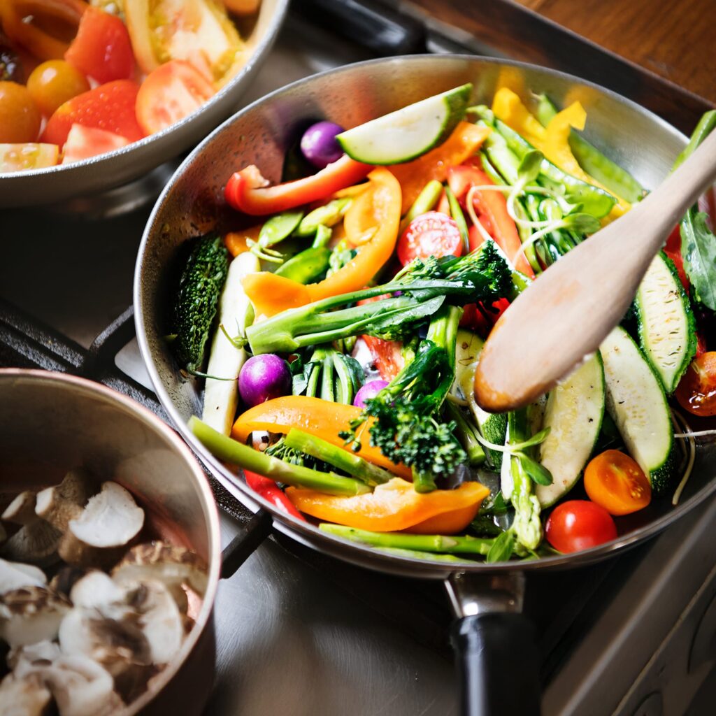 Colorful fresh vegetables, including broccoli, bell peppers, cherry tomatoes, and zucchini, are being sautéed in a pan with a wooden spoon on a stovetop. Nearby pots contain mushrooms and sliced tomatoes.