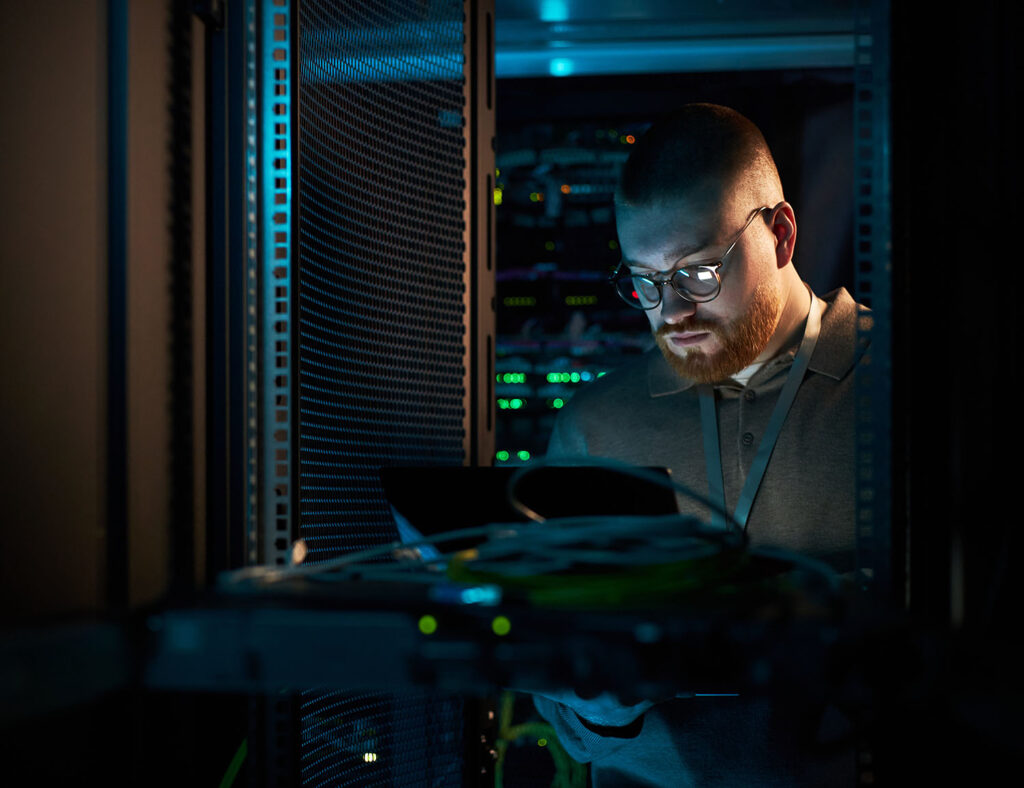 A man with glasses and a beard works on a laptop in a dimly lit server room, surrounded by server racks with blinking lights and cables.