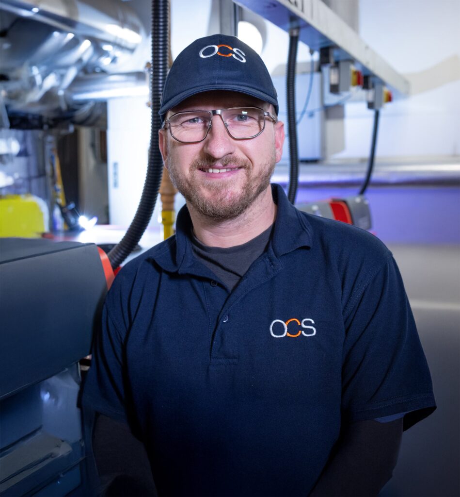 A smiling man wearing glasses, a navy blue OCS cap, and a matching OCS polo shirt stands in an industrial or factory setting with machinery in the background.