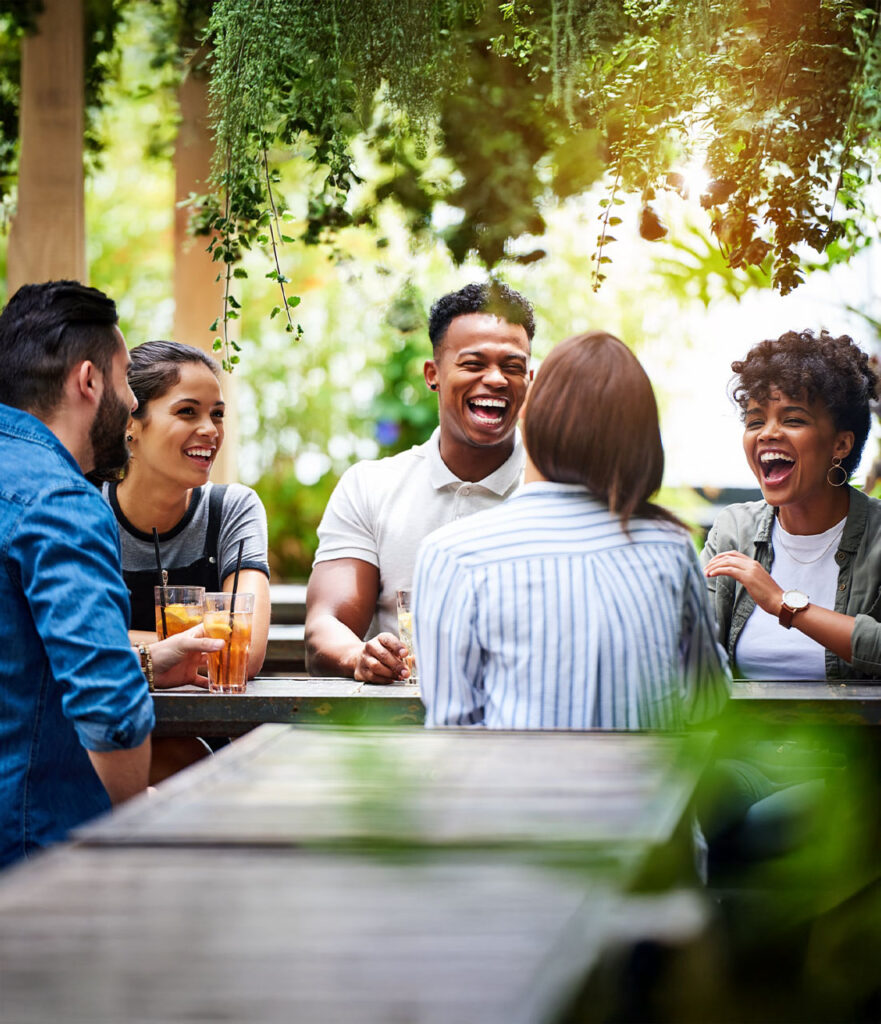 Five friends sit at an outdoor table surrounded by greenery, laughing and enjoying drinks together. The atmosphere is bright and lively, with sunlight streaming through the leaves above.