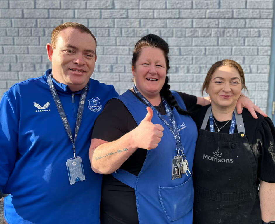 Three people wearing uniforms and name badges stand smiling together in front of a brick wall. The person in the middle gives a thumbs-up, and the person on the right wears a black apron with Morrisons on it.
