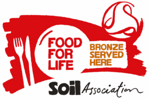 Red and white logo with a fork and knife, the words “FOOD FOR LIFE” in a circle, “BRONZE SERVED HERE” in a gold box, and “Soil Association” in black script below.