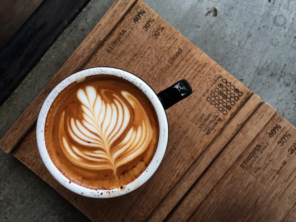A cup of cappuccino with leaf-shaped latte art sits on a wooden surface next to a menu featuring different coffee origins and details.