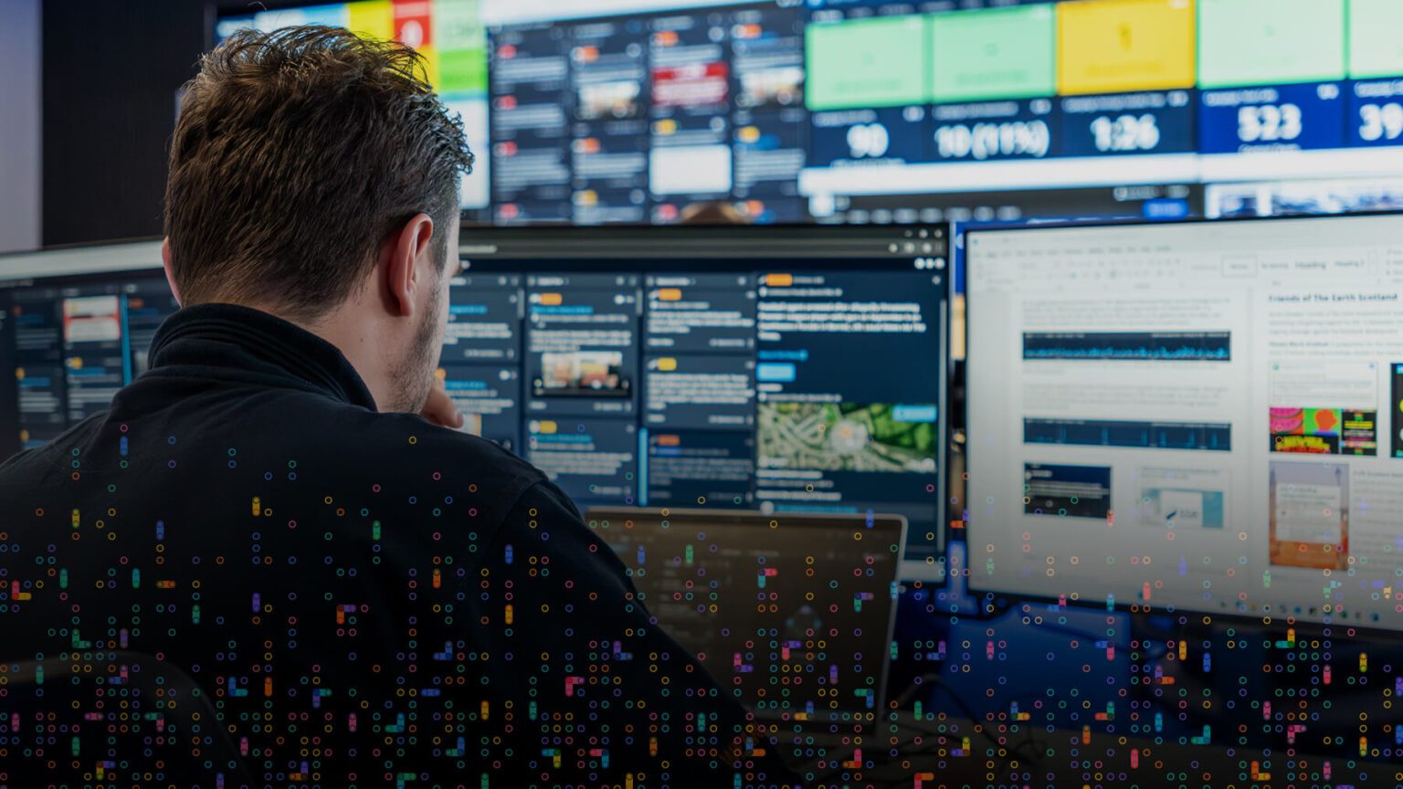 A person sits at a desk, facing multiple computer monitors displaying data, graphs, and news feeds, with a colorful overlay of digital dots and a large screen wall in the background.
