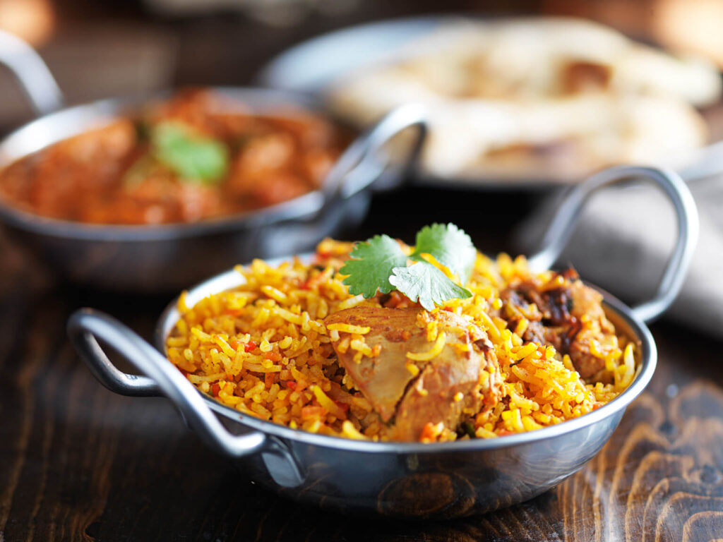 A metal bowl filled with biryani rice and pieces of meat, garnished with fresh cilantro, sits on a wooden table. In the background, another dish and pieces of naan bread are visible, slightly out of focus.