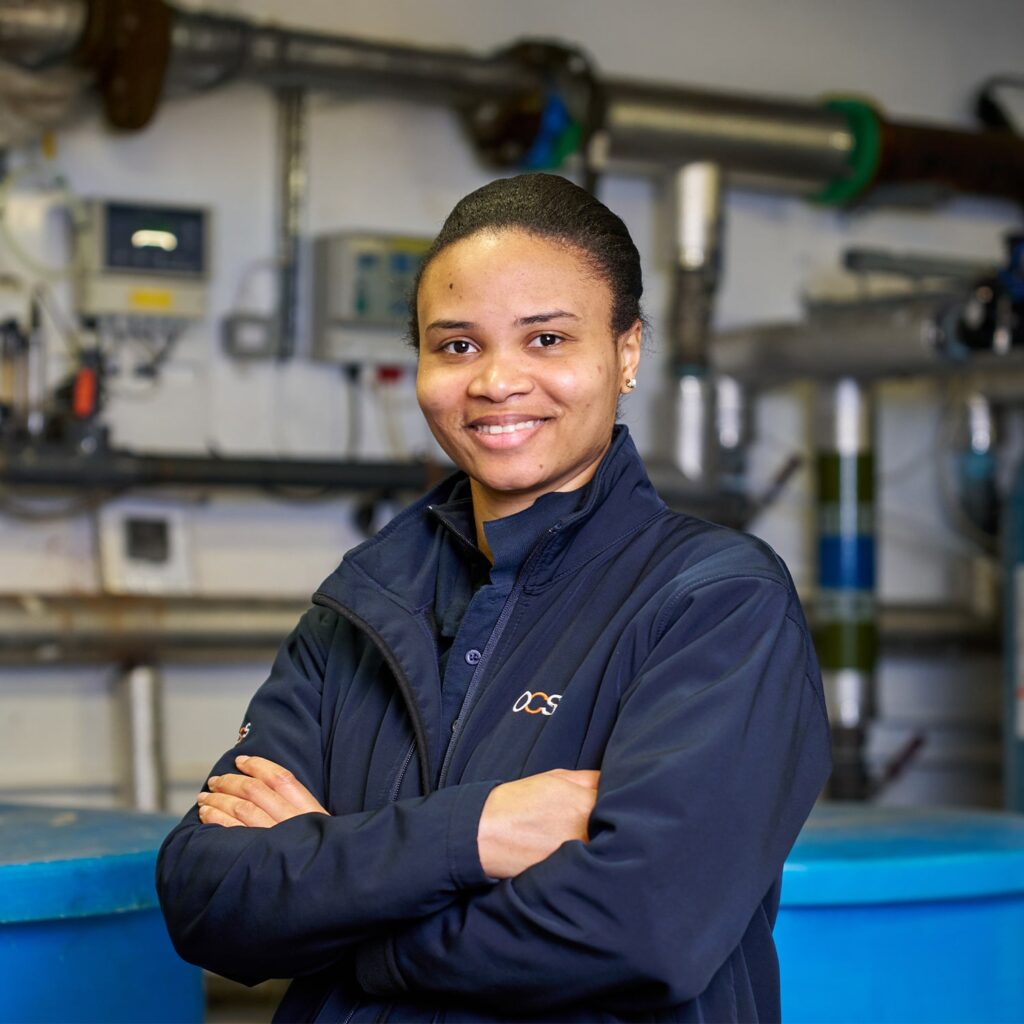 A person wearing a dark jacket stands with arms crossed and smiles at the camera in an industrial setting with pipes, gauges, and control panels in the background.