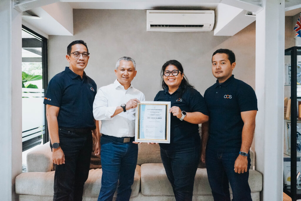 Four people stand indoors, three in matching navy shirts with OCS logos, one in a white shirt. The woman in the group holds a framed certificate, and they all smile at the camera. A couch and air conditioner are visible behind them.