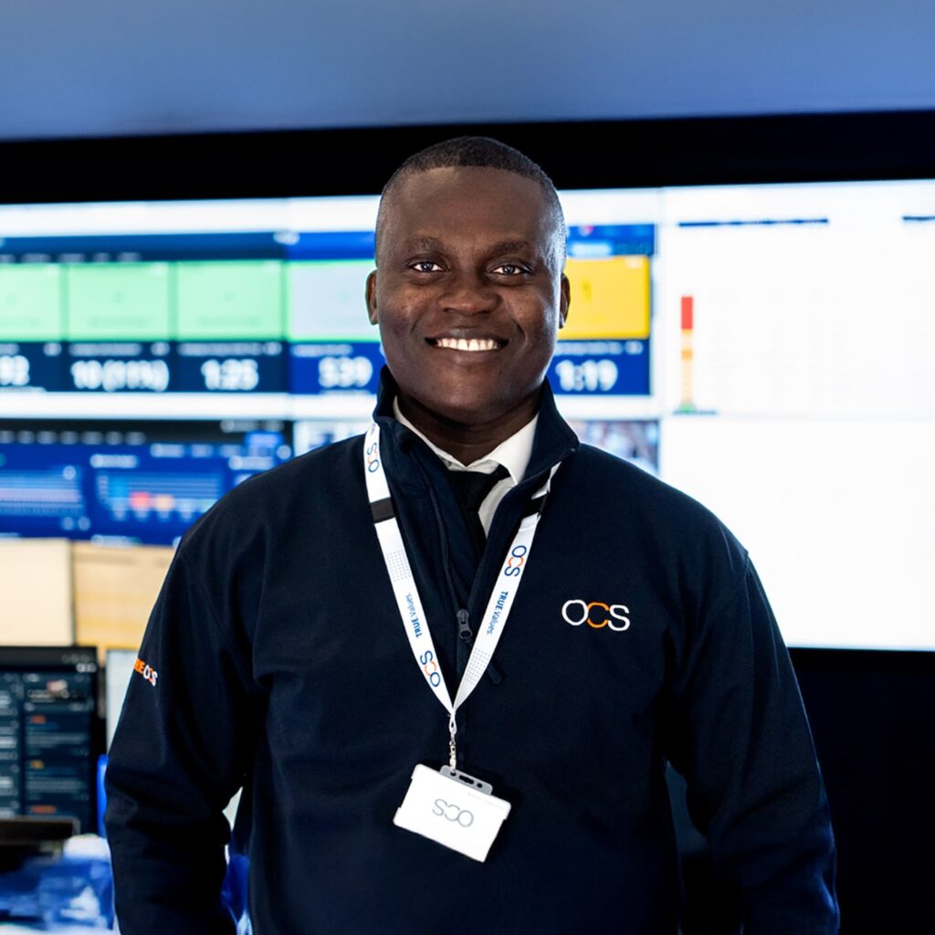 A man wearing an OCS-branded jacket and lanyard smiles while standing in front of large computer screens displaying various data and charts in a modern control room.
