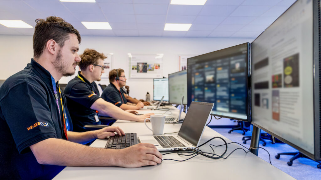 Four people are seated at a row of desks in an office, each working on a laptop with large monitors. The focus is on the person in the foreground typing, while others concentrate on their screens.