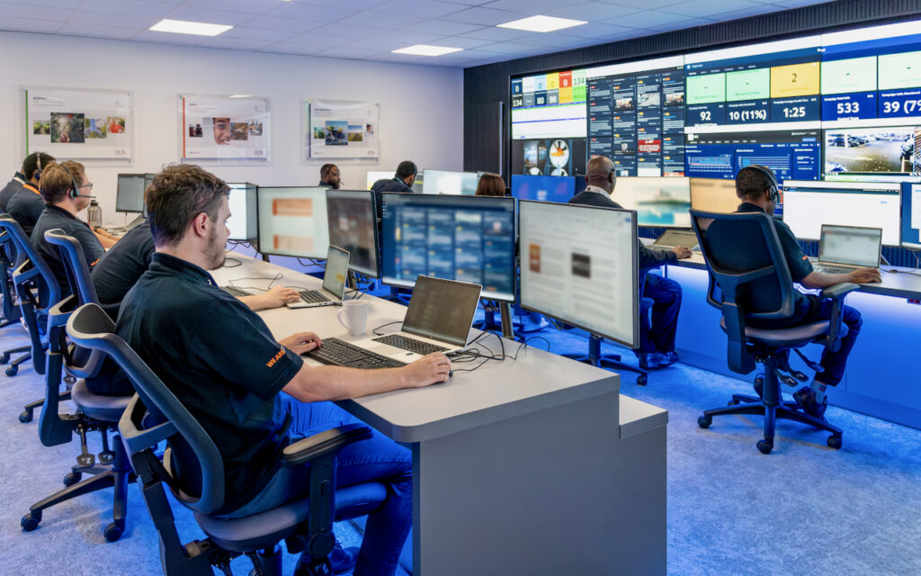 A modern control room with multiple people working at computer desks, large monitors displaying data and dashboards on the front wall, and several smaller screens on each workstation.
