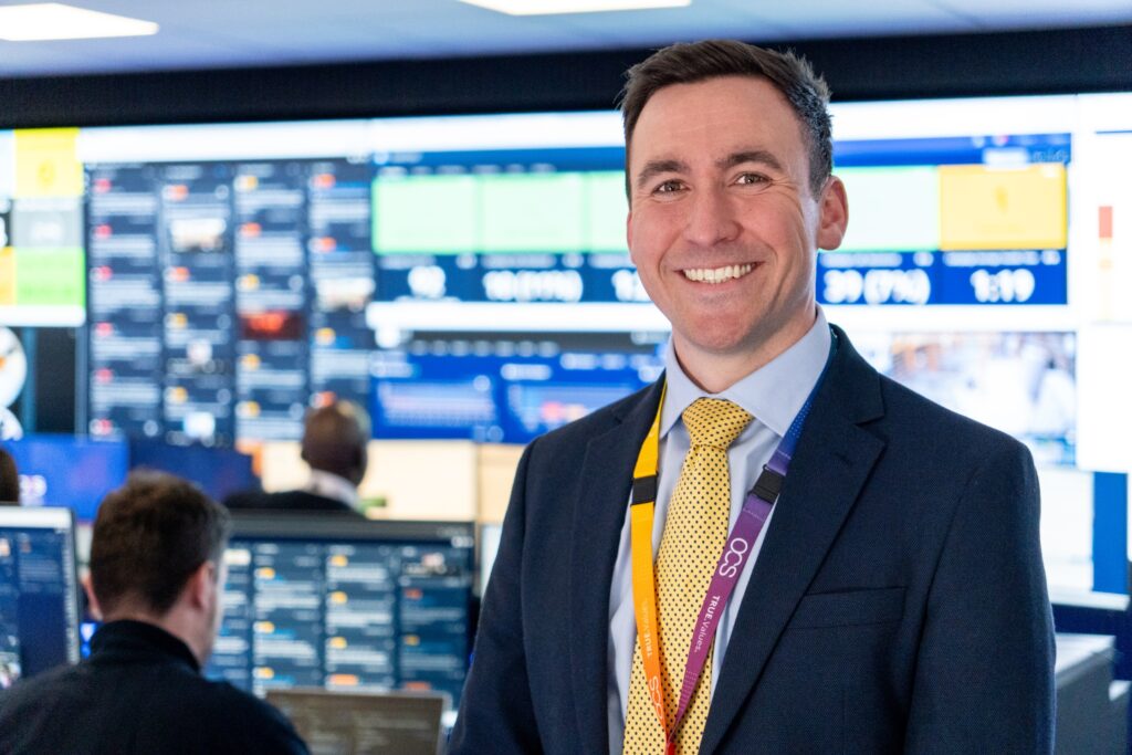 A man in a suit and yellow tie smiles at the camera in a control room with multiple monitors and people working at computers in the background.