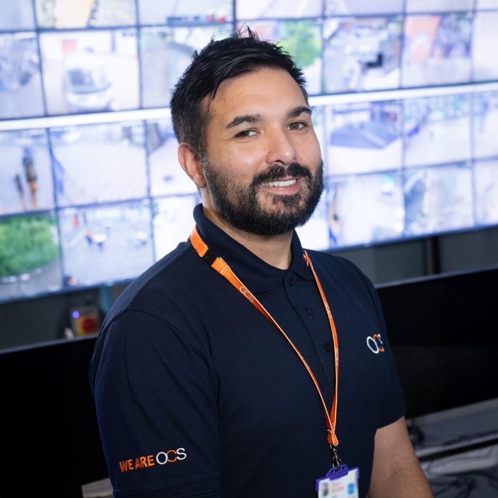 A man with a beard wearing a navy blue WE ARE OCS polo shirt and an orange lanyard stands in front of a wall of surveillance monitors, smiling at the camera.