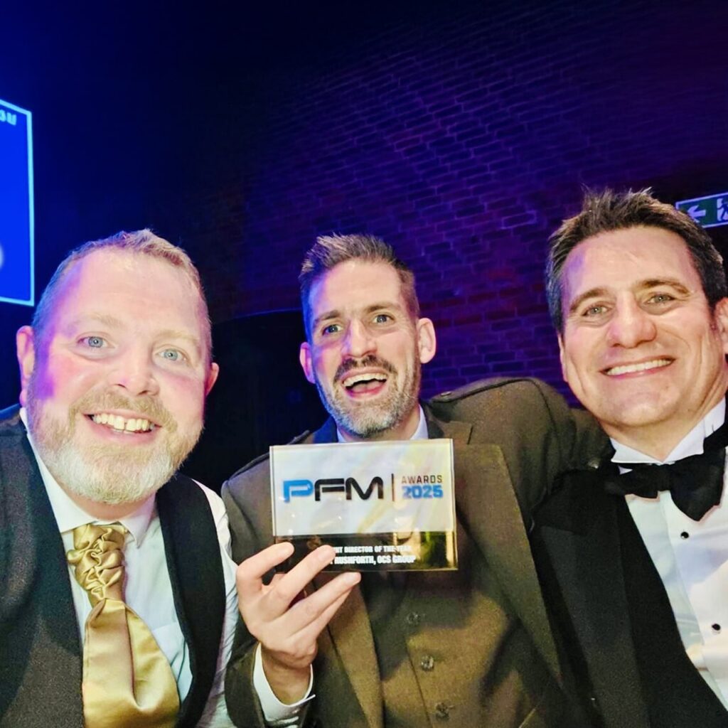 Three smiling men in formal wear pose for a selfie at an event. The man in the center holds a glass award that reads PFM Awards 2023. The background is dark with brick walls and event lighting.