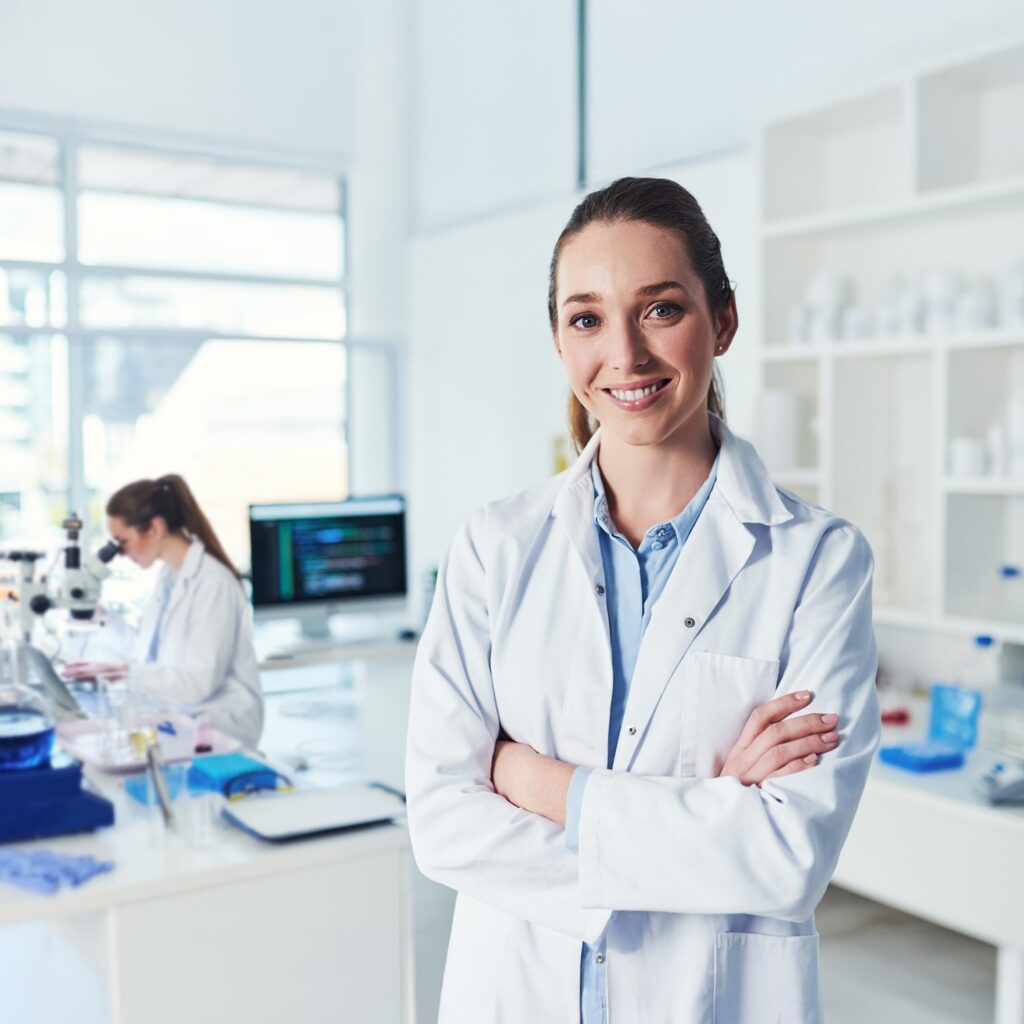A woman in a white lab coat stands smiling with arms crossed in a bright, modern laboratory, while another person works at a lab bench in the background.