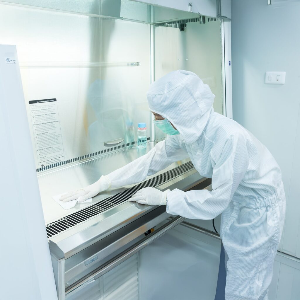 A scientist wearing full-body protective clothing and gloves works inside a sterile laboratory, wiping down the surface of a biosafety cabinet with a cloth for cleanliness and contamination control.