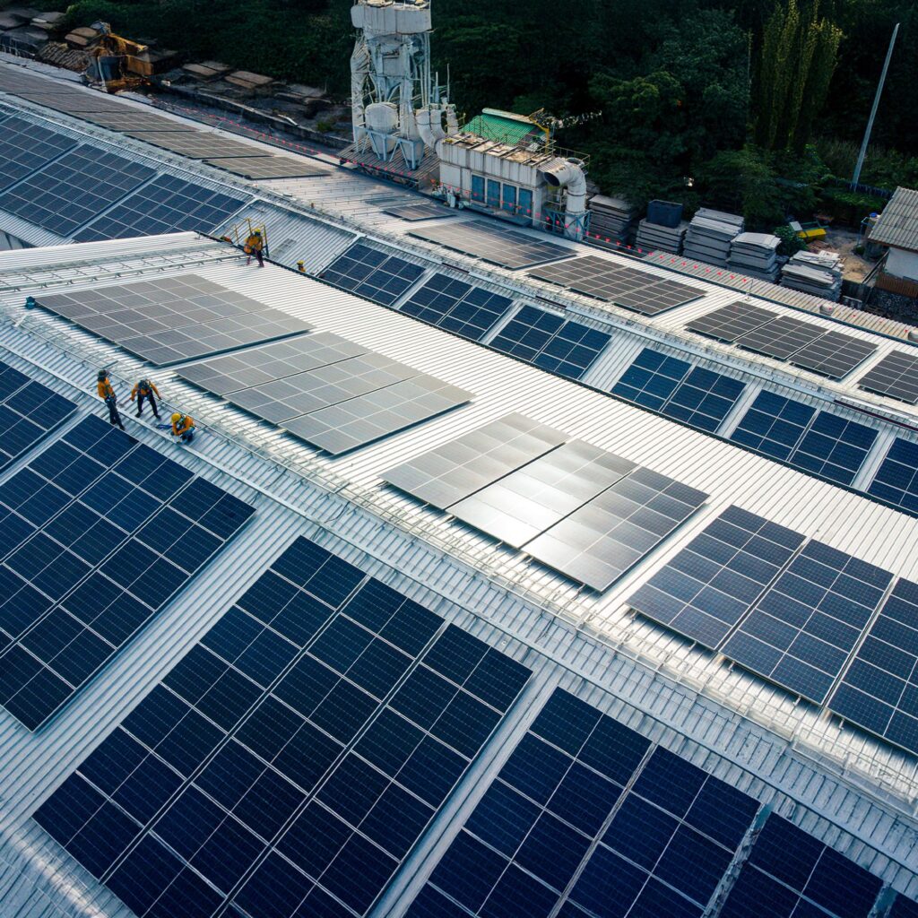 Aerial view of workers installing solar panels on the roof of a large industrial building, surrounded by rows of existing solar panels and nearby warehouses.