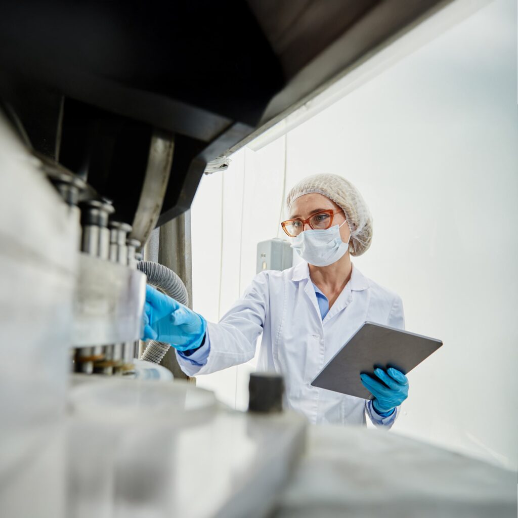 A scientist wearing a lab coat, hairnet, mask, and gloves operates laboratory equipment while holding a tablet, working in a clean, bright lab environment.