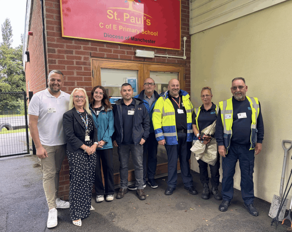 A group of eight adults, some in high-visibility jackets, stand smiling in front of the entrance to St. Pauls C of E Primary School, beneath a red sign, outdoors on a paved area.