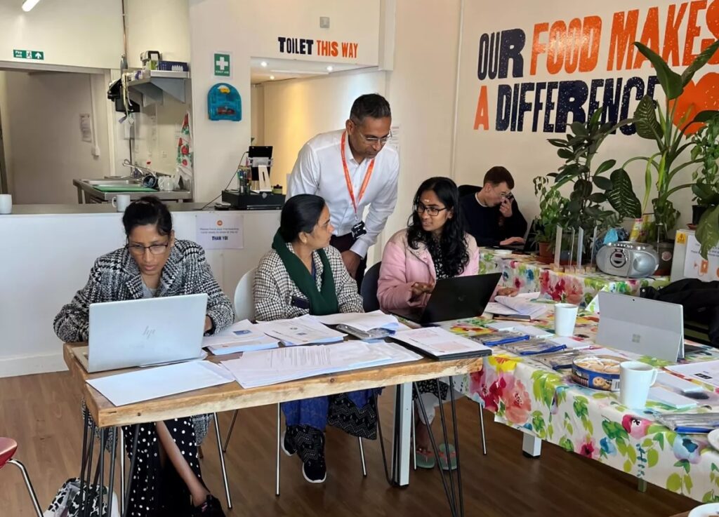 Four people sit at a table with laptops and papers, while one person stands behind them. The room has colorful decor and a sign on the wall reads “OUR FOOD MAKES A DIFFERENCE.” A kitchen is visible in the background.