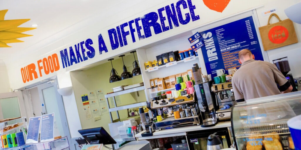 Bright café interior with a sign reading “OUR FOOD MAKES A DIFFERENCE” above the counter. A person stands behind the counter near colorful mugs, a coffee machine, and a menu board on the wall.