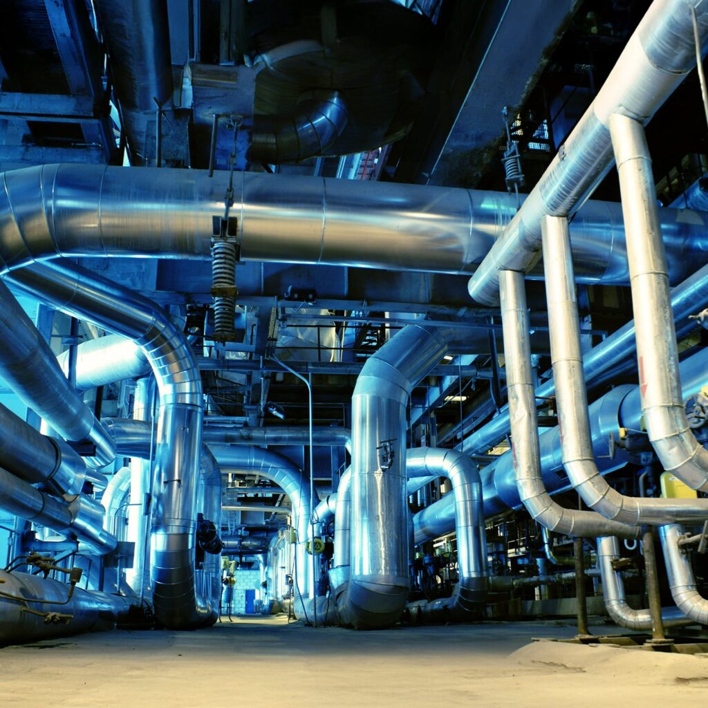 Large network of shiny metal pipes and ducts in an industrial facility, with blue-toned lighting and various support beams and machinery visible in the background.