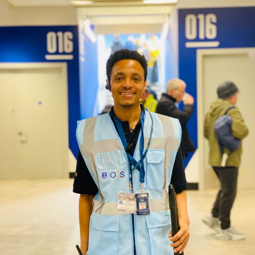 A smiling security staff member wearing a light blue reflective vest and ID badge stands in a hallway between two doors labeled 016; people walk in the background.