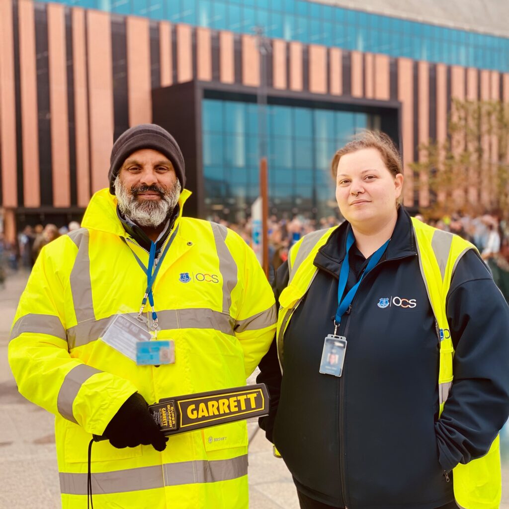 Two security staff wearing OCS uniforms and ID badges stand outside a modern building. One holds a handheld metal detector labeled GARRETT. Both are looking at the camera. Other people are visible in the background.
