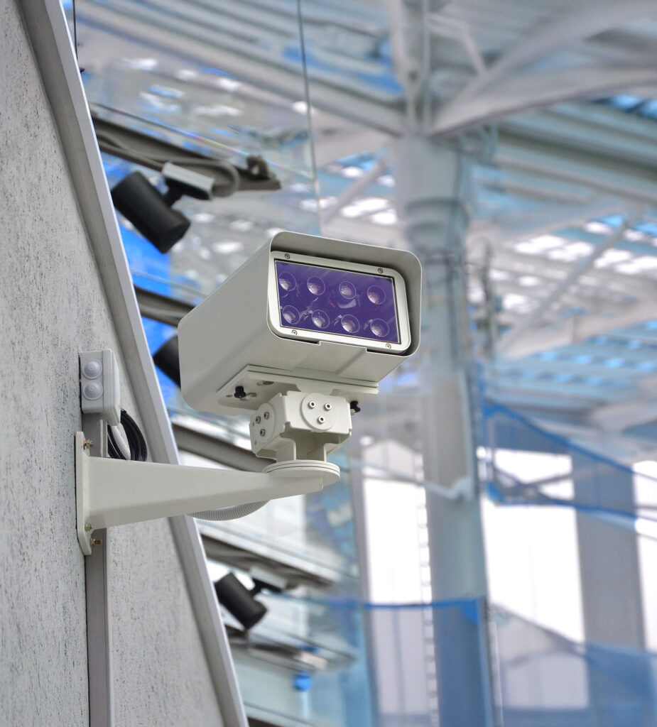 A close-up of a modern security camera mounted on a wall indoors, with a blurred background of large windows and metal beams.