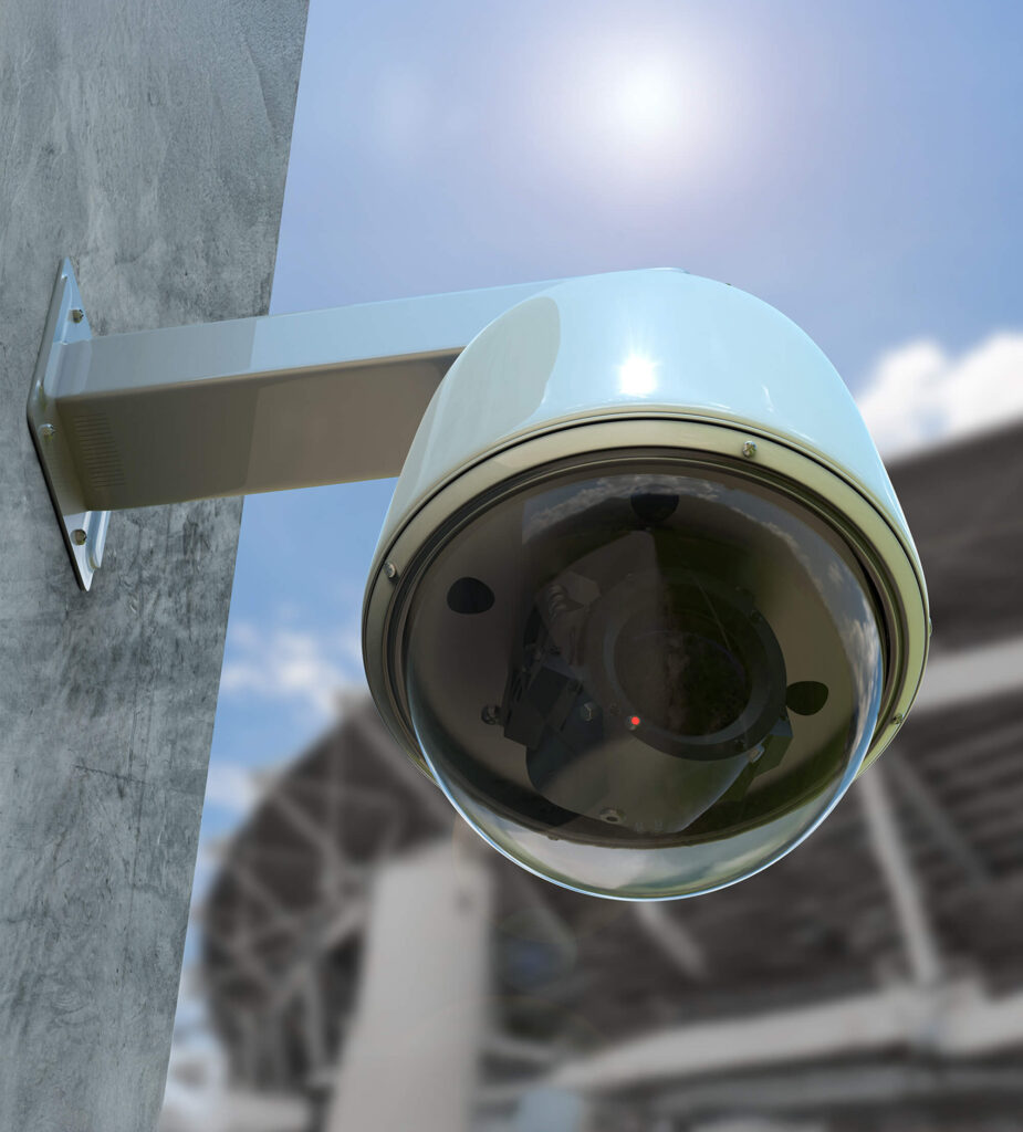 A dome security camera mounted on a wall outdoors, with a blurred background showing part of a building structure and a bright sun in the sky.
