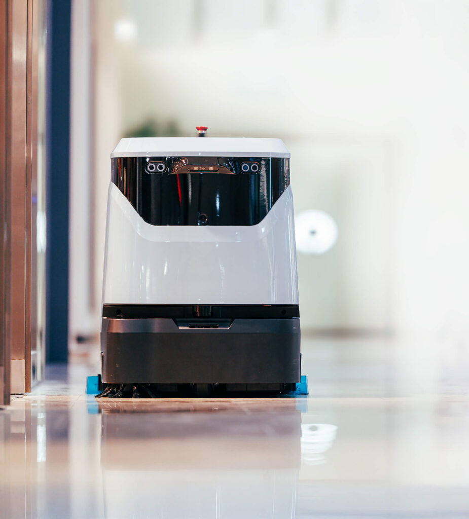 A white, box-shaped floor-cleaning robot operates in a modern, brightly lit hallway with reflective floors and blurred background details.
