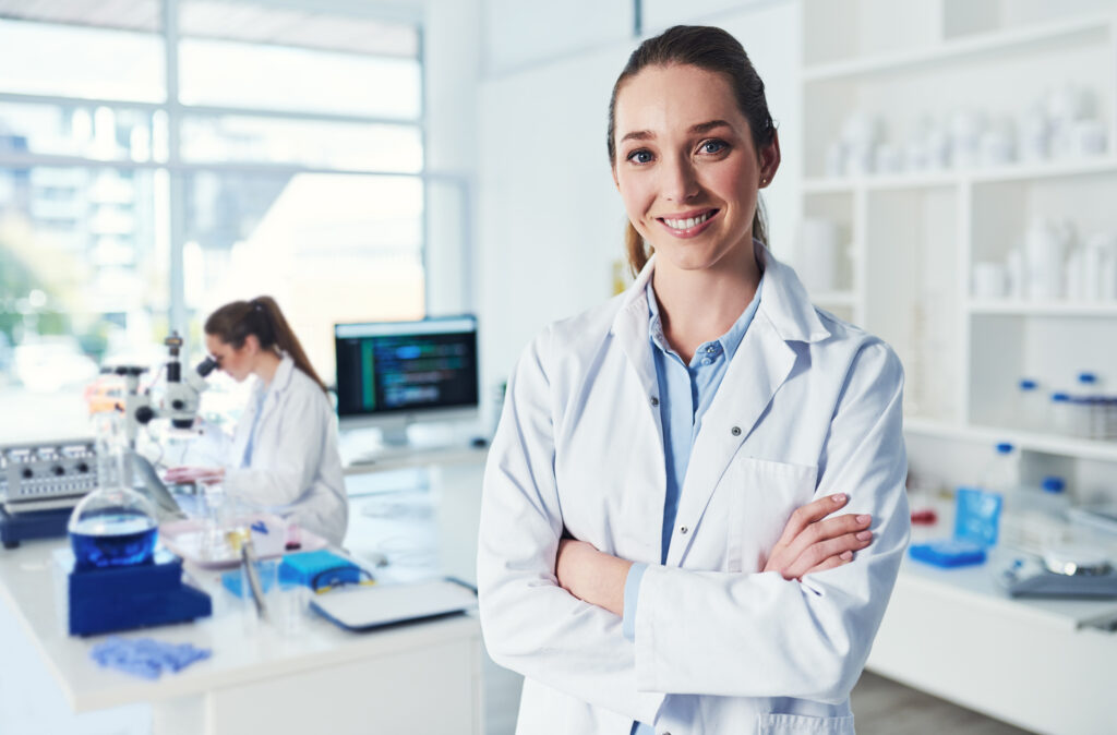 A smiling scientist in a white lab coat stands with arms crossed in a bright laboratory, while another scientist works at a desk with lab equipment and a microscope in the background.