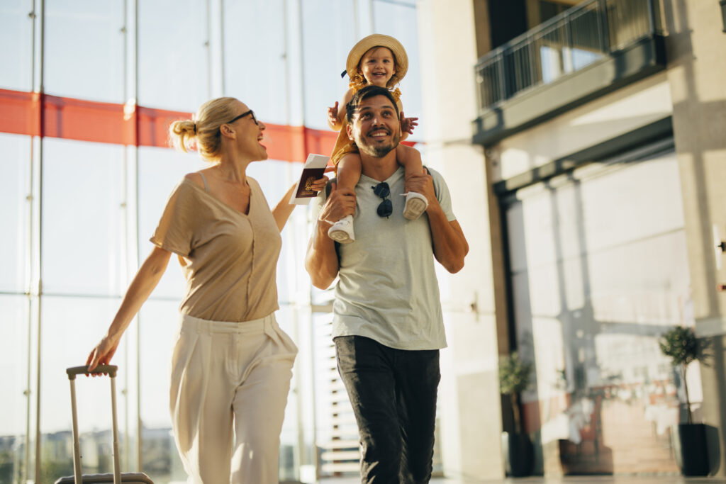 A smiling family walks through a bright airport terminal. An adult carries a suitcase, another adult carries a child on their shoulders. The child is wearing a hat and smiling happily. Sunlight streams in through large windows.