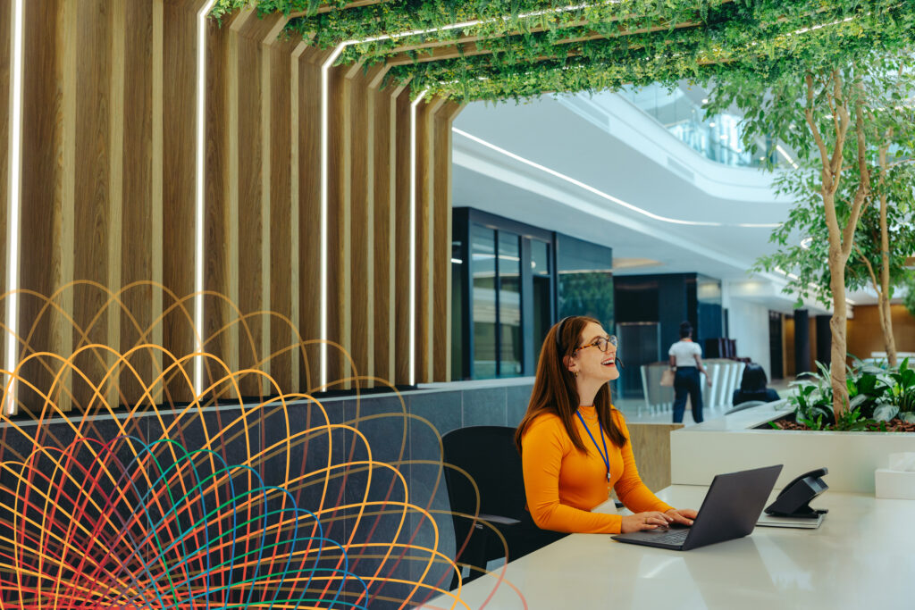 A woman in an orange top sits at a modern office desk with a laptop, smiling. The workspace features green plants, wooden accents, and a colorful sculpture in the foreground.