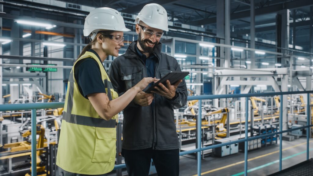 Two factory workers wearing hard hats and safety vests stand on an indoor balcony, smiling and looking at a tablet device, with industrial machinery and equipment visible in the background.