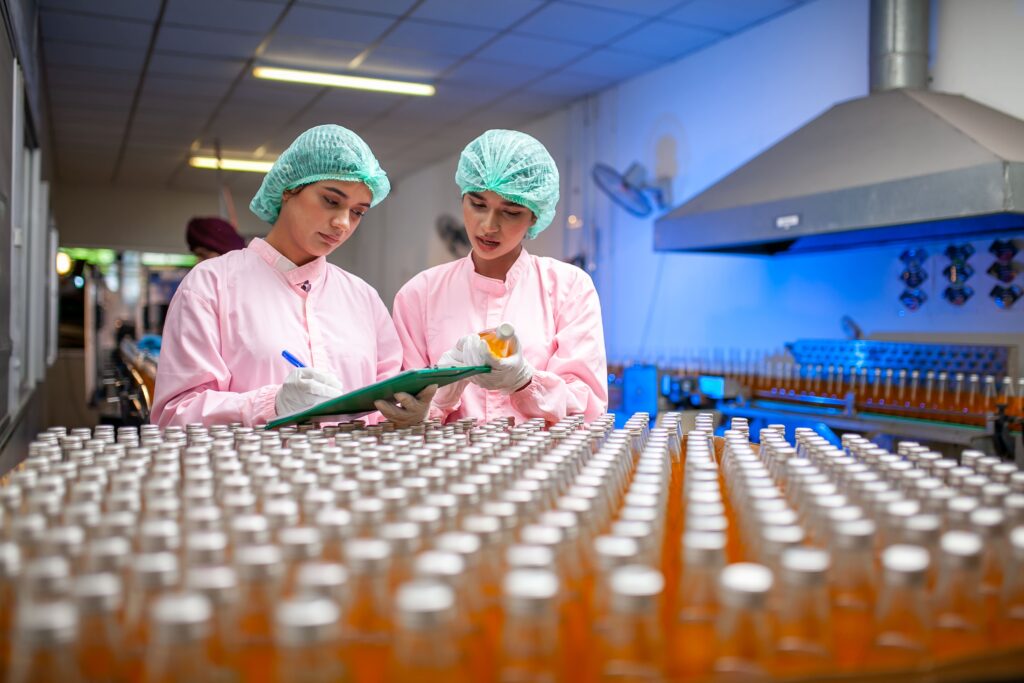 Two workers in pink uniforms and hairnets inspect and take notes in a beverage factory, standing beside rows of bottled drinks on a production line.