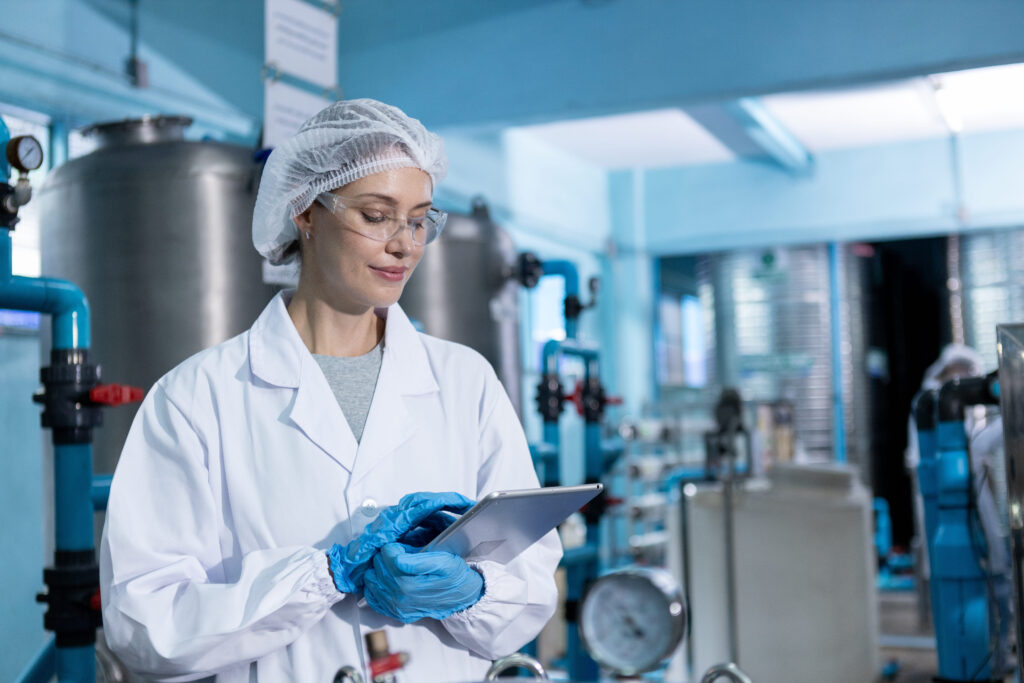 A scientist wearing a lab coat, hairnet, gloves, and safety glasses uses a tablet in a laboratory or industrial facility with large metal tanks and blue pipes in the background.