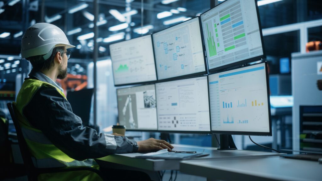 A person wearing a hard hat and high-visibility vest sits at a desk, monitoring multiple screens displaying graphs, charts, and data in a modern industrial control room.