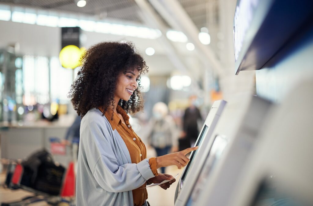 A woman with curly hair smiles while using a touchscreen kiosk at an airport terminal, with suitcases and other travelers visible in the background.