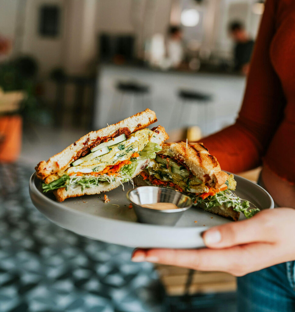 A person holds a plate with a sliced veggie sandwich filled with lettuce, tomato, cucumber, and sprouts, and a small cup of dipping sauce. The background shows a blurred café setting.
