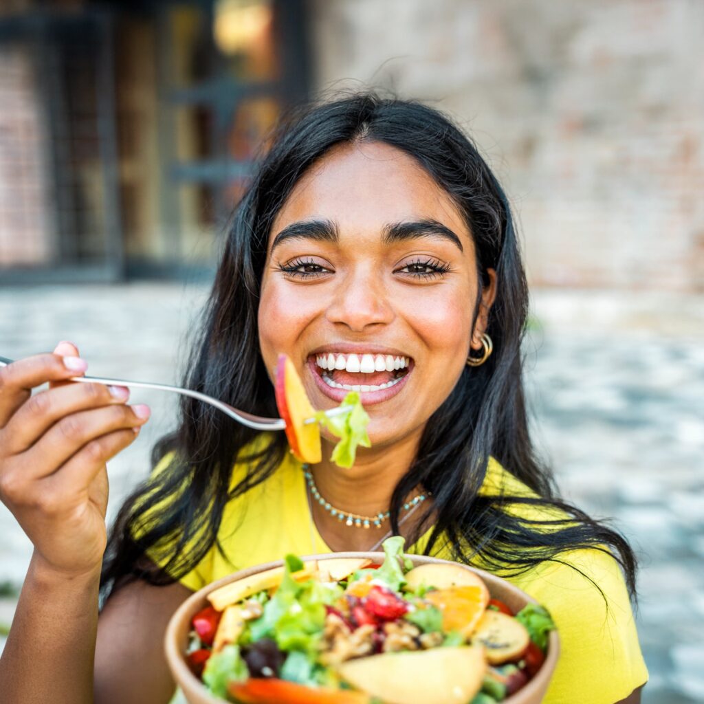 A smiling woman with long dark hair holds a bowl of salad and lifts a fork with a bite of food toward her mouth. She is outdoors, wearing a yellow shirt, with a blurred background behind her.