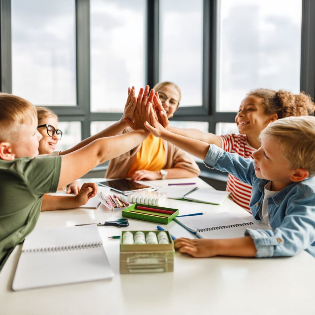 A group of happy children sitting at a table in a classroom high-fiving each other, with notebooks and school supplies in front of them and a smiling teacher in the background.
