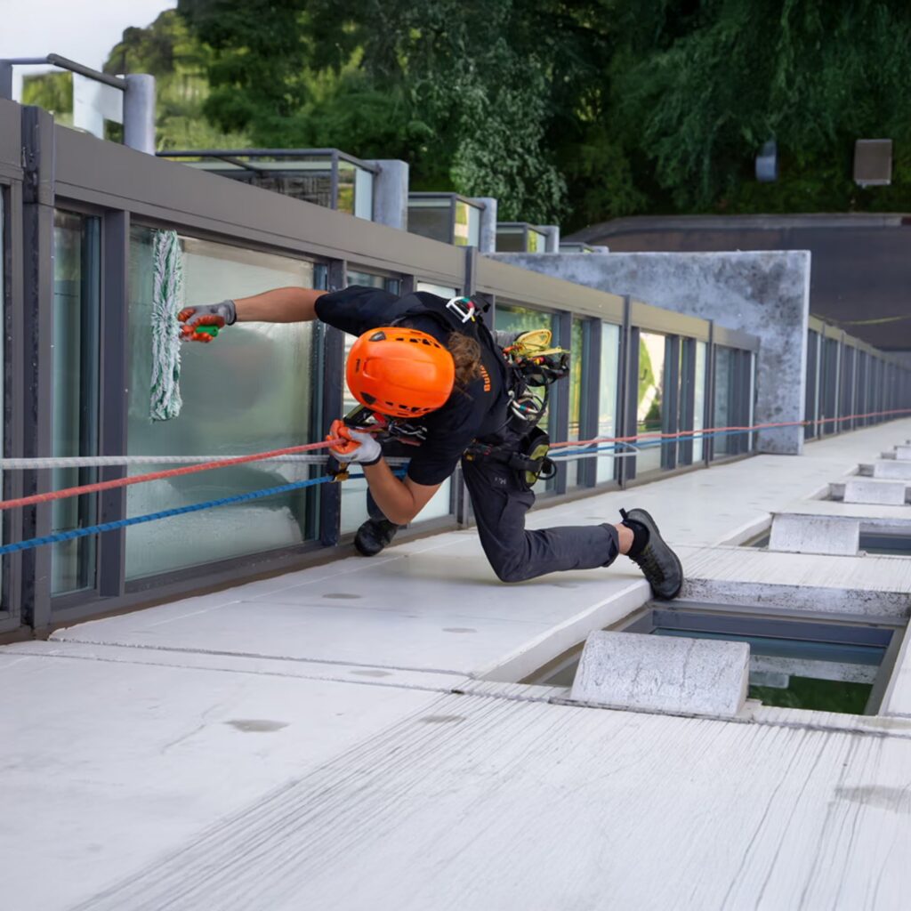 A worker wearing a helmet and safety harness cleans windows on the exterior of a building while secured with ropes, demonstrating industrial rope access techniques. Trees and a road are visible in the background.