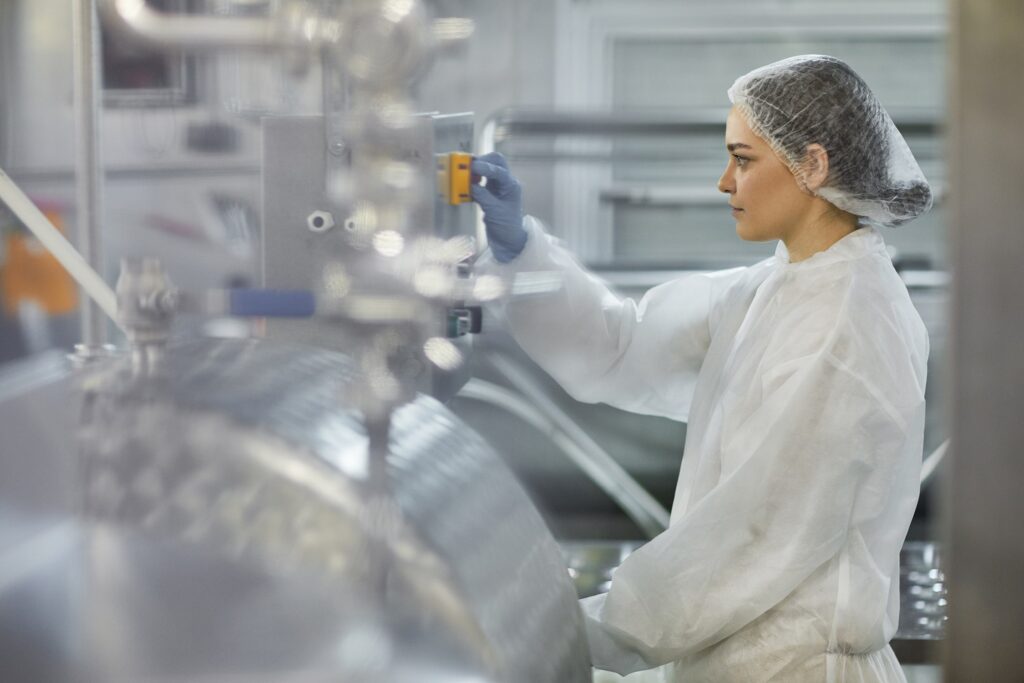 A woman in protective clothing and a hairnet operates machinery in a clean, industrial facility, likely a food or pharmaceutical processing plant. She is focused on adjusting equipment controls.