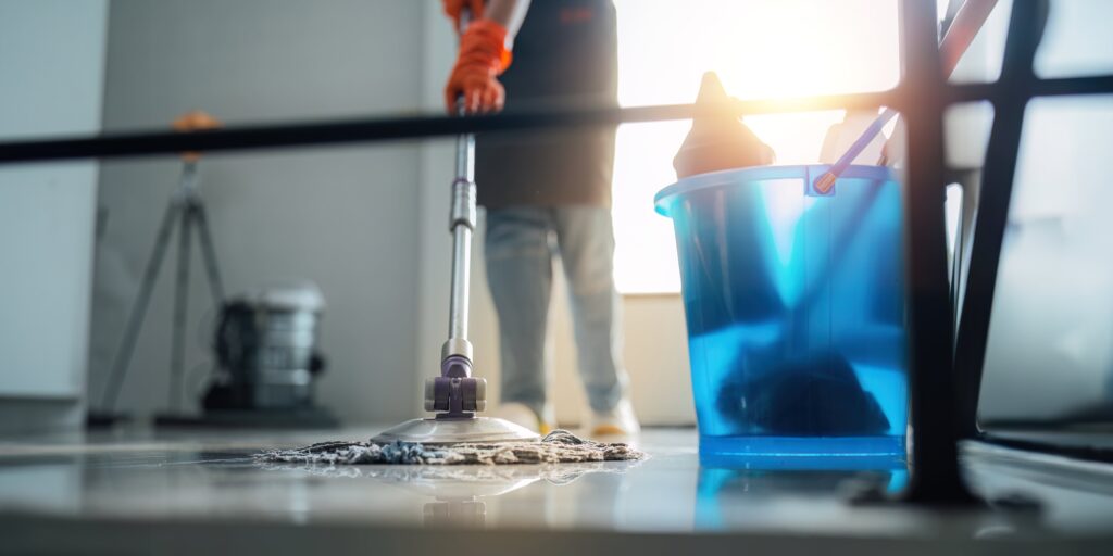 A person wearing orange gloves mops a shiny floor near a blue bucket filled with cleaning supplies, with sunlight streaming in from a window in the background.